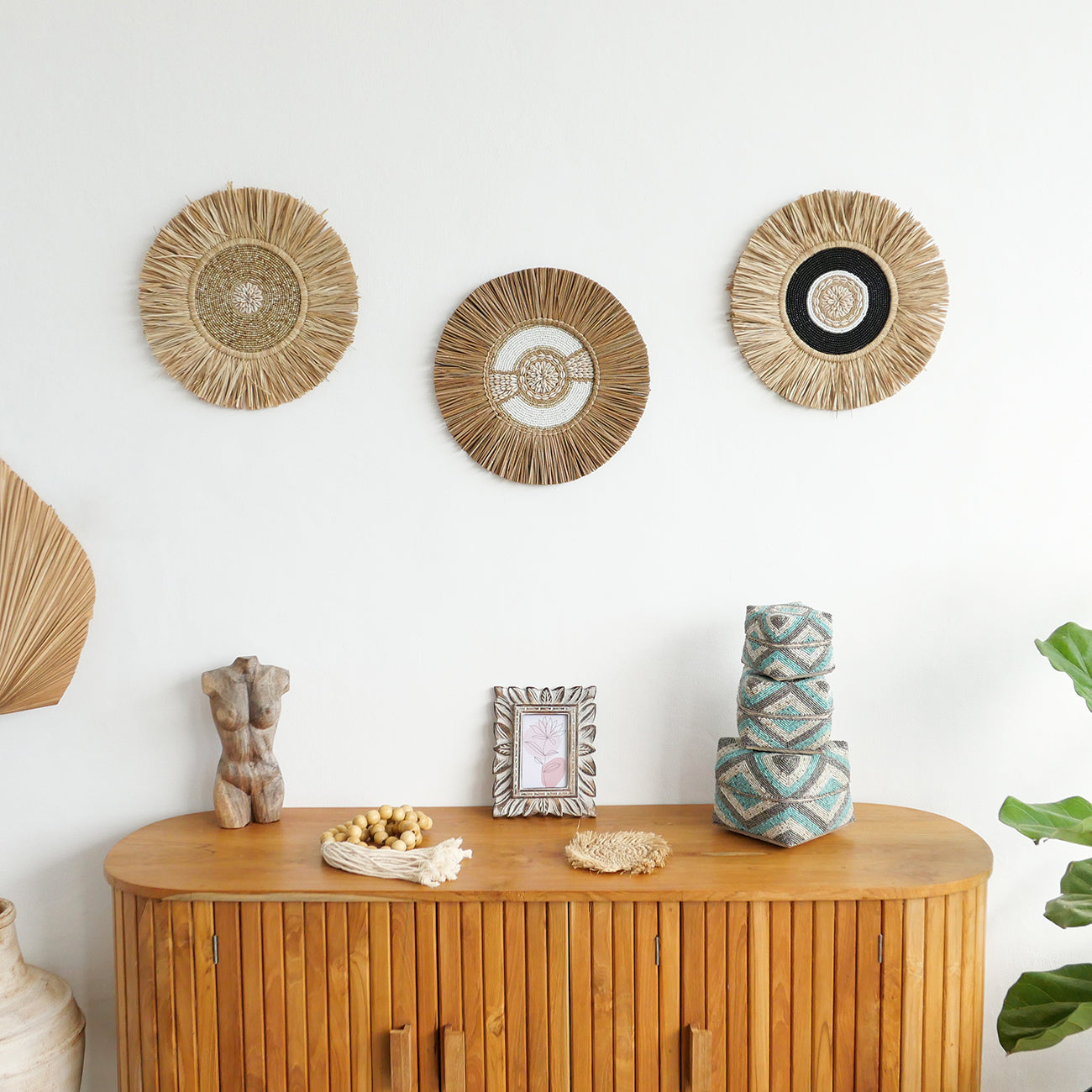 A set of three circular wall decor items made from natural materials, hanging on a wooden wall above a wooden cabinet with decorative items and a framed picture.